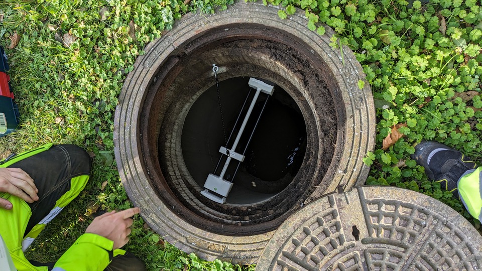 A sensor is mounted across an open storm drain; a hand, knee and foot are visible against the surrounding greenery.